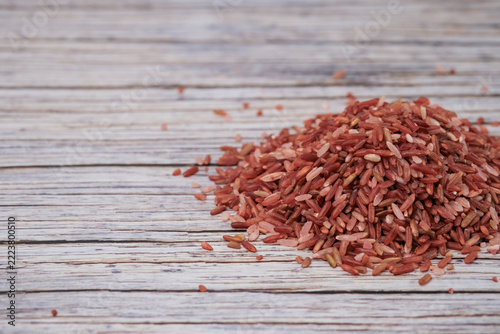 Red rice grains pile on weathered wooden surface. Texture contrasts between grain and wood add visual interest. Natural, rustic setting evokes wholesome, earthy qualities. Ideal for food, health
