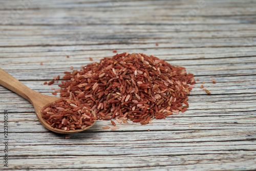 Red rice grains spill from a wooden spoon. Rice rests on weathered, textured wood surface. Natural tones highlight wholesome, earthy quality. Ideal for health, nutrition, or culinary themes