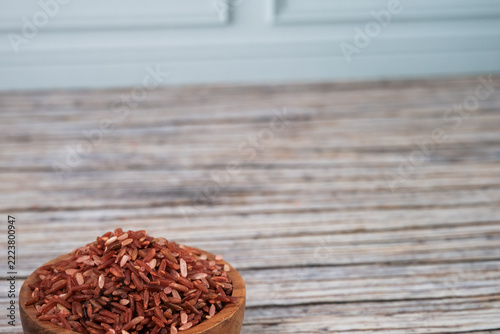Red rice grains fill a small wooden bowl. Bowl rests on weathered wooden planks. Natural tones evoke rustic, wholesome appeal. Image suggests organic, earthy, healthy food. Suitable for nutrition