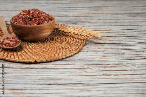 Rice grains fill a wooden bowl with earthy tones. Woven mat holds bowl and spoon beside wheat stalks. Wooden chopsticks rest nearby, adding rustic charm. Soft light highlights textures