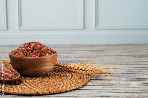 Rice grains fill a wooden bowl with earthy tones. Woven mat holds bowl and spoon beside wheat stalks. Wooden chopsticks rest nearby, adding rustic charm. Soft light highlights textures