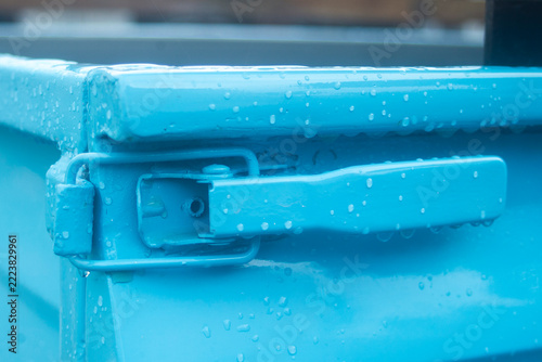 Close up of a blue metal truck bed tailgate latch covered in raindrops.
A high-quality close-up photograph focusing on a heavy-duty toggle latch mechanism on the tailgate corner of a vintage pickup 