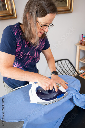 Mature woman holding fabric taut while ironing a blue shirt at home.