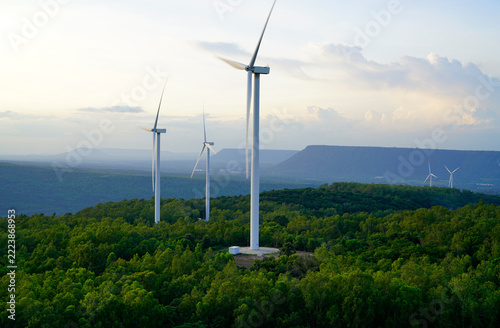 Wind turbines generating renewable electricity across a green forest and mountain landscape at sunset. Sustainable energy, clean technology, low carbon development and environmental conservation.