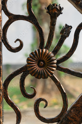 wrought iron gate. Metalwork blooms with intricate curls and aged patina. Sunlight glints softly through the wrought design. Background hints at garden or courtyard setting. Ideal for rustic, vintage