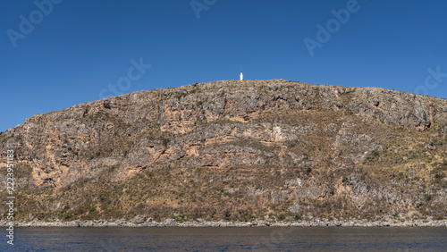 A high-altitude navigable lake. On the coastal hill, a white lighthouse against a blue sky. Scant vegetation on the slopes. In the foreground, the surface of the water. Peru. Lake Titicaca. 
 