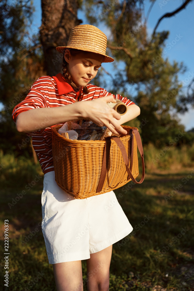 Fototapeta premium A young woman lifestyle portrait holding a wicker basket during a picnic in the park. The girl looks at organic food items with soft sunset sunlight. Leisure time activity.