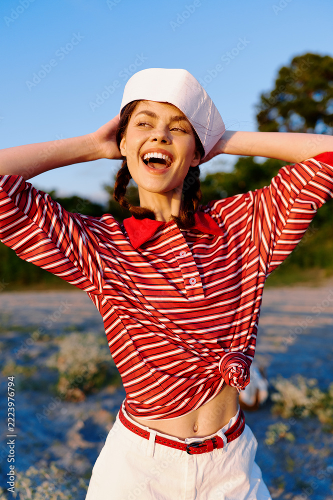 Naklejka premium Cheerful young woman wearing sailor hat and striped shirt laughs while standing on sandy beach outdoors. Authentic lifestyle portrait capturing pure joy during summer day.