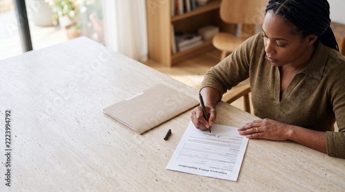 Young african american woman signing insurance policy application form at stone table. Adult applicant completing official legal contract paperwork during personal finance management.
