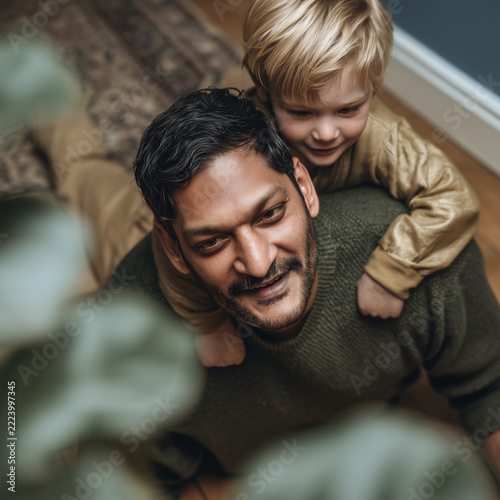 Man and young boy share a quiet, affectionate moment. The boy rests happily on the man's shoulders. Both smile softly, eyes filled with warmth and joy. They lie on a cozy, patterned rug beside a plant