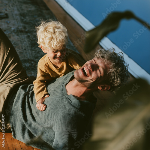 Sunlight bathes a joyful father and child. Child sits on father's chest, smiling wide. Father laughs, eyes closed, arms open. They rest on wooden floor, near a window