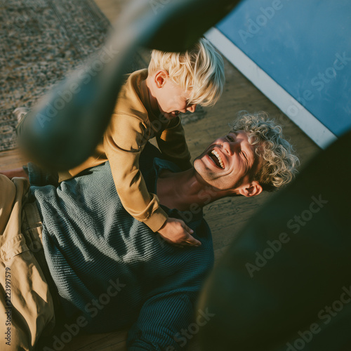 Sunlight bathes a joyful father and child. Golden curls frame a laughing, relaxed face. Child hugs father, eyes closed in delight. Warm tones evoke cozy, intimate moments. Perfect for parenting