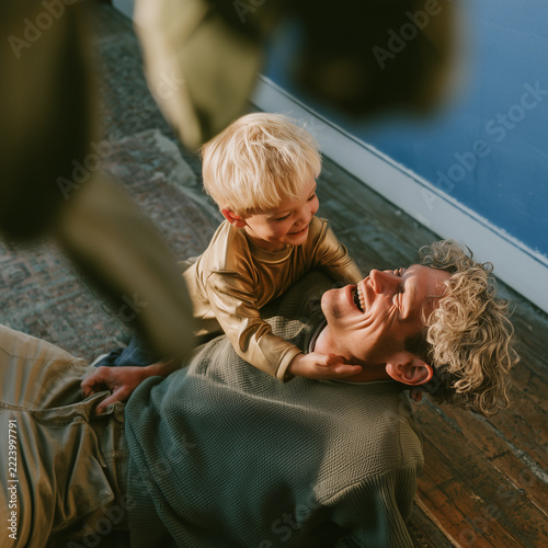 Golden light bathes a joyful father and child. Child hugs father with bright, laughing eyes. Father smiles wide, eyes closed in pure delight. They lie on wooden floor, sharing warmth and joy