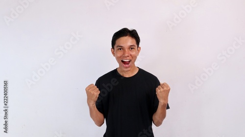 Excited young Asian man shouting for joy while pumping both clenched fists in a victory celebration against a white studio background.