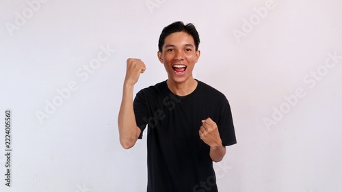 Triumphant young Asian man laughing cheerfully while raising his right fist in the air to celebrate victory against a white background.
