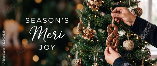 Hand hanging Maori wooden ornament on Christmas tree background with warm holiday bokeh lights and festive text for New Zealand Pacific island Season's Meri Kirihimete celebration banner template.
