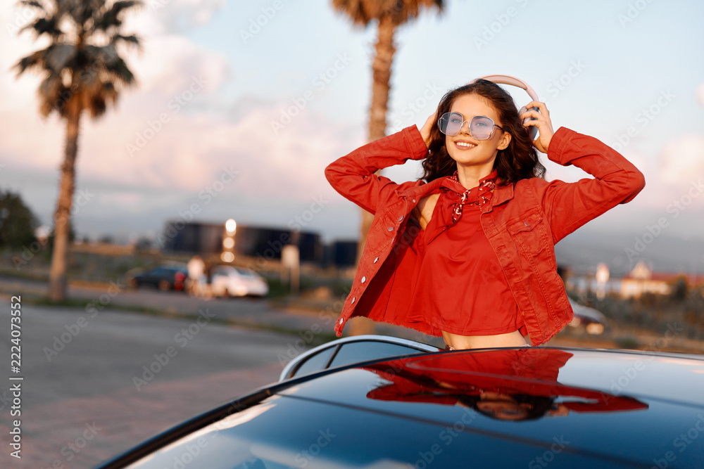 Fototapeta premium A stylish young woman in red jacket and sunglasses stands near a black car during sunset. Concept for travel, freedom, and modern lifestyle on the road.