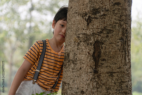 Shy young Asian boy wearing a striped shirt and suspenders, hiding and peeking from behind a large tree trunk in an outdoor park, expressing childhood emotion, playing hide and seek in nature