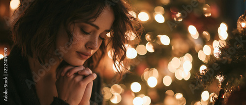 A serene woman with her hands clasped in prayer, eyes closed in contemplation, surrounded by the soft glow of Christmas lights