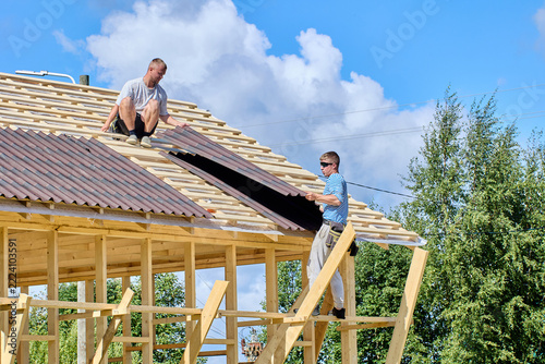 Asphalt roofing sheets being positioned by two workers on wooden roof battens during installation of roofing on timber structure.