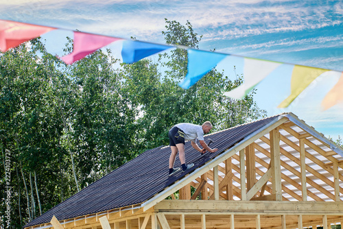 Corrugated bitumen sheets are fixed by worker using nails and hammer on roof frame during construction of wooden building.