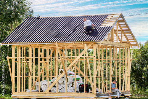 Asphalt roofing panels covering most of pitched roof as worker installs roofing above timber frame with exposed studs and diagonal braces.