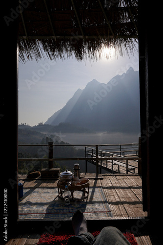  Traveler enjoys breakfast inside a cozy bamboo house framing the misty mountain view of Doi Luang Chiang Dao in Chiang Mai Thailand for a peaceful nature getaway. Vertical photo