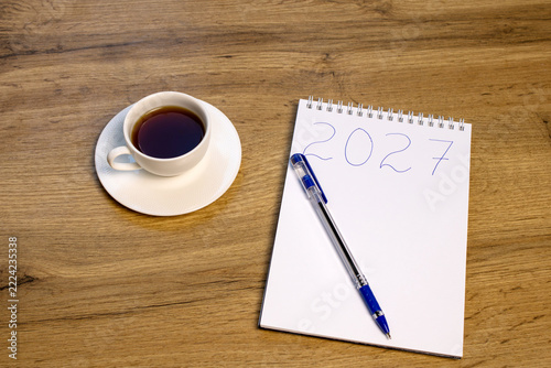 A white notepad marked 2027 with a blue pen and a cup of black coffee on a wooden desk. New year planning and personal goal setting concept.