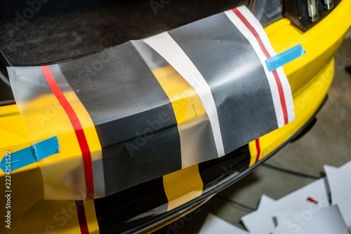 Partially applied racing stripes on the rear bumper of a yellow sports car during vinyl wrap installation in an automotive workshop