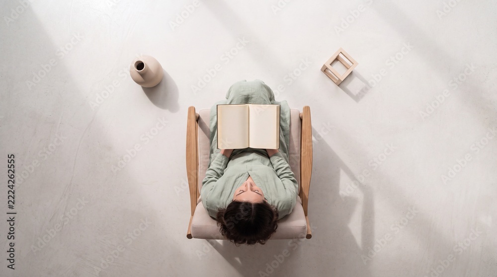Naklejka premium Top down overhead view of a young woman sitting in a minimalist wooden armchair reading an open book in a bright, clean indoor space with soft natural lighting and long artistic shadows