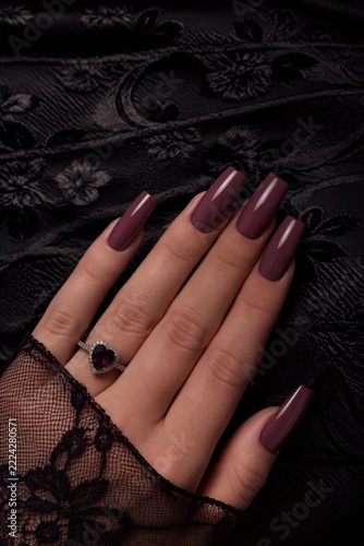 Close-up of a woman's hand with polished burgundy nails, adorned with a heart-shaped ruby ring, resting on a black lace floral fabric background.