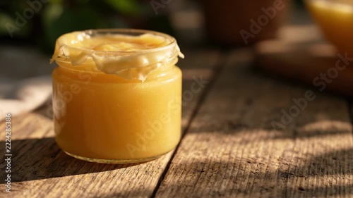 A clear jar filled with golden honey sits on a weathered wooden surface, illuminated by soft sunlight filtering through green foliage. Nature’s essence radiates in this tranquil scene.
