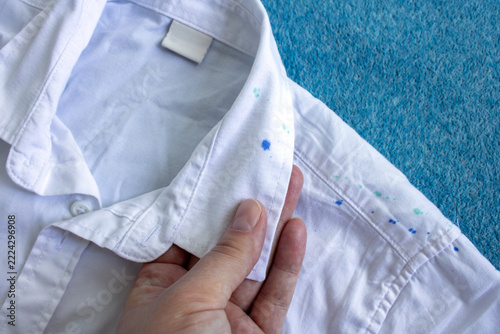 A hand holds the collar of a white shirt next to noticeable blue watercolor stains on the fabric. A concept illustrating clothing damage and stain removal during washing.