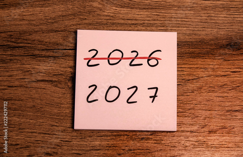 Pink sticky note with 2026 crossed out and 2027 written below on a wooden surface. New year transition and fresh start concept.