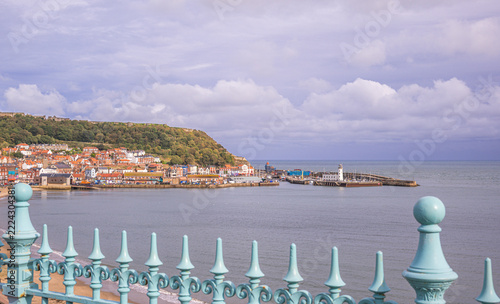 A seaside town with a harbour and lighthouse and railings are in the foreground.