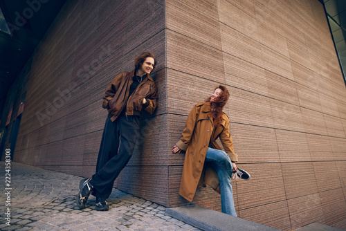 Fashionable young couple smiling at each other while leaning against building corner on cobbled street. City break travel, coordinated outfits concept.