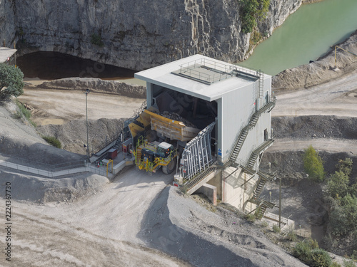 Aerial view of a large dump truck unloading rocks into a cement crushing facility at a quarry, surrounded by rocky terrain and a small pond in the United Kingdom.