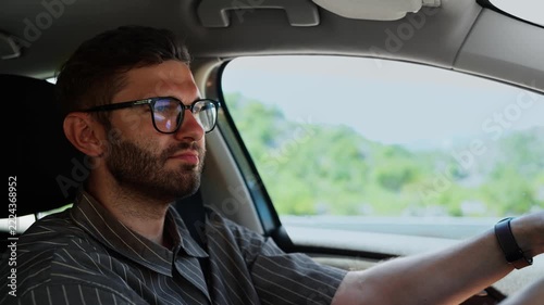 Focused young man wearing glasses driving a car through sunny green countryside, concentrating on the road ahead while enjoying a calm relaxed solo journey