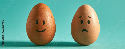 Two brown eggs with happy and sad faces drawn on them stand on teal surface, expressing contrasting emotions in simple, creative concept