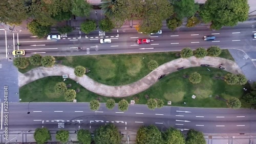 Aerial drone footage of a curved pedestrian walkway surrounded by green lawns and palm trees between busy urban roads. Top down view of moving vehicles, traffic lanes and city infrastructure.