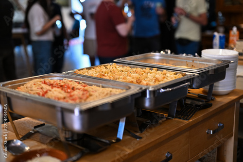 Pasta dishes in chafing dishes at a catering buffet with guests in the background