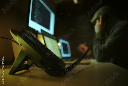 Defocused Hacker in Dark Hoodie Making Telephone Call Among Computer Screens. Telephone Fraud Concept.