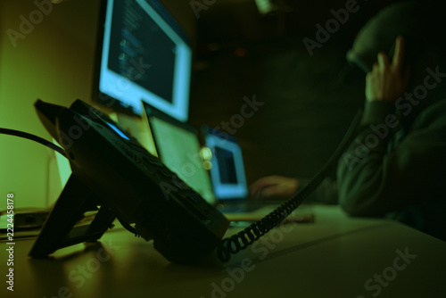 Defocused Hacker in Dark Hoodie Making Telephone Call Among Computer Screens. Telephone Fraud Concept.