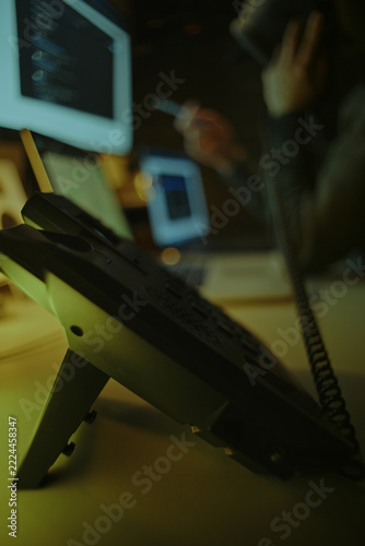 Defocused Hacker in Dark Hoodie Making Telephone Call Among Computer Screens. Telephone Fraud Concept.