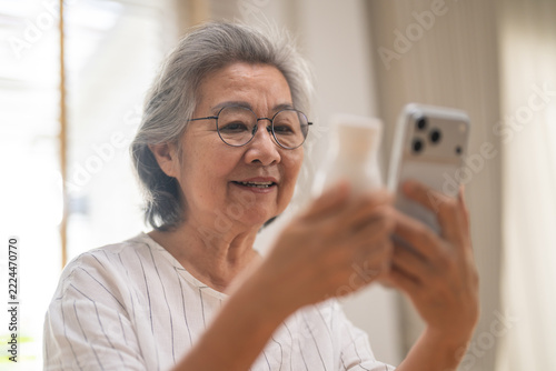 Healthcare e-commerce and e-pharmacy digital technology business concept, Asian elderly woman using smartphone and holding medicine bottle at home, Smiling senior checking online pharmacy application