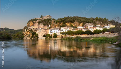   A picturesque historic village with an ancient fortress nestled atop a steep rocky cliff, perfectly reflected in the calm waters of a wide river under a bright blue sky  