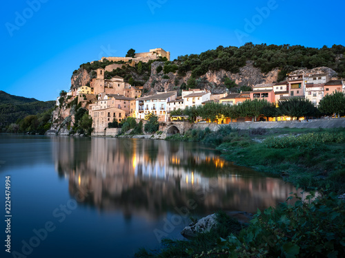   A picturesque historic village with an ancient fortress nestled atop a steep rocky cliff, perfectly reflected in the calm waters of a wide river under a bright blue sky  