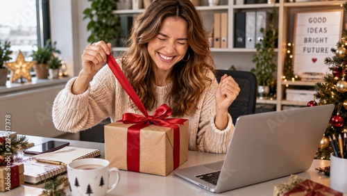 A happy woman opens a kraft gift tied with red ribbon beside her laptop at work