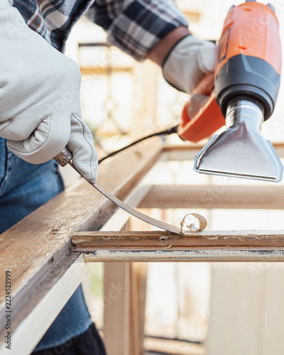 Adult carpenter wearing protective leather gloves, using a heat gun and a wood scraper to remove paint from an old wooden window. Construction industry, carpentry. Restoration. 