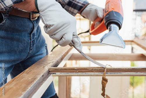 Adult carpenter wearing protective leather gloves, using a heat gun and a wood scraper to remove paint from an old wooden window. Construction industry, carpentry. Restoration. 